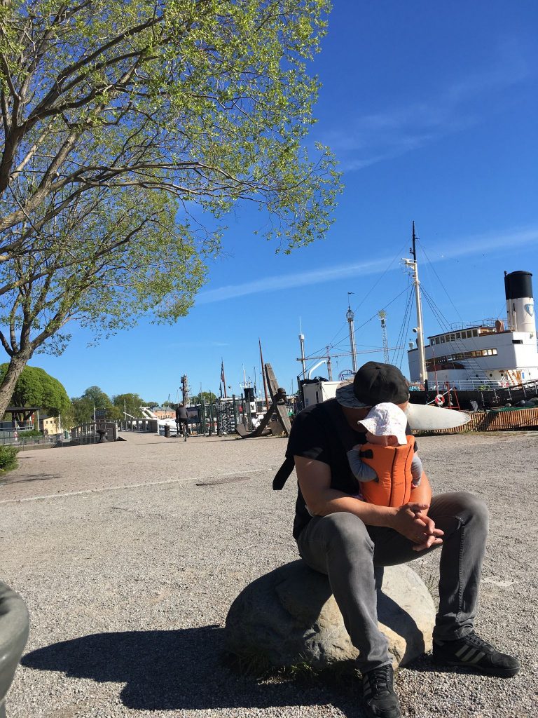Mann mit Baby in Trage sitzt an Hafen in Stockholm. Podcast Thema